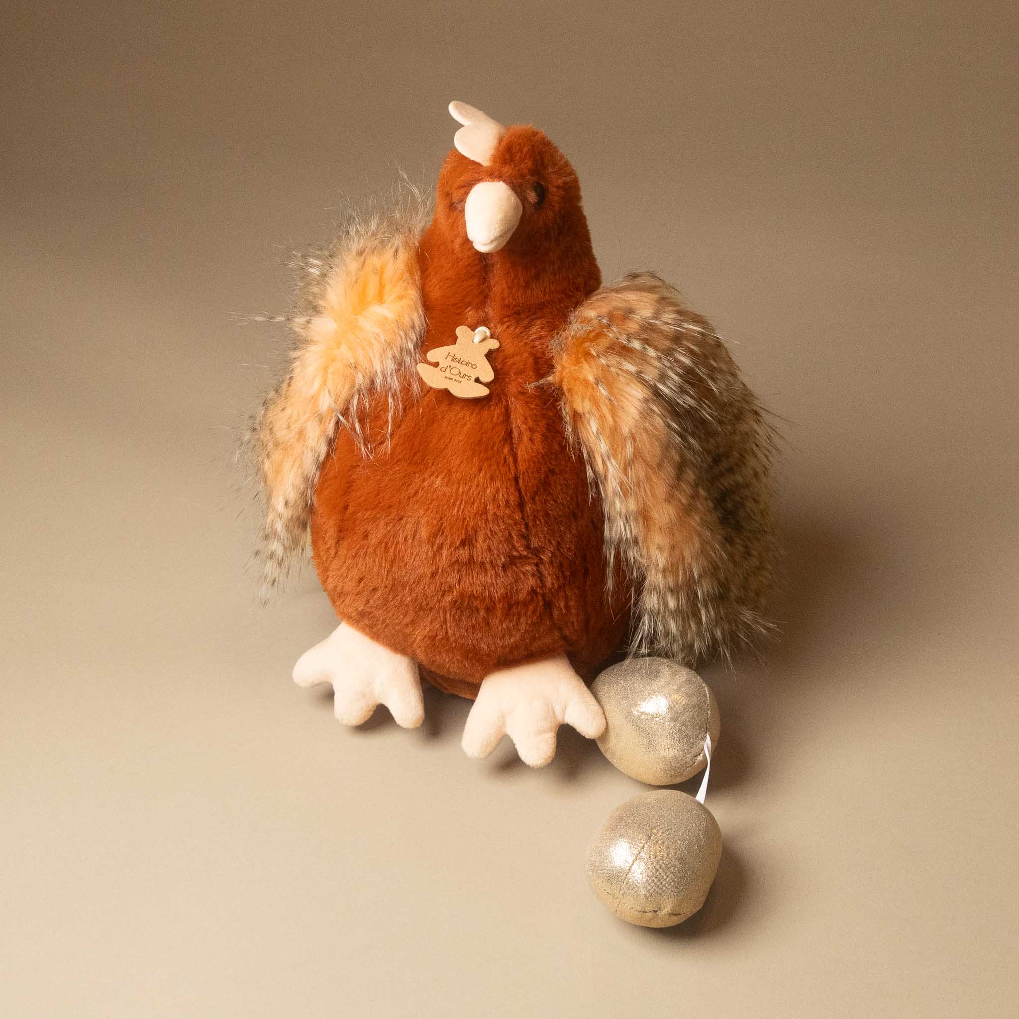 A plush floofy-chicken--her-golden-eggs--rouge with brown and beige feathers, white beak and feet, and two silver hanging weights attached to its tail, photographed on a plain beige studio background.