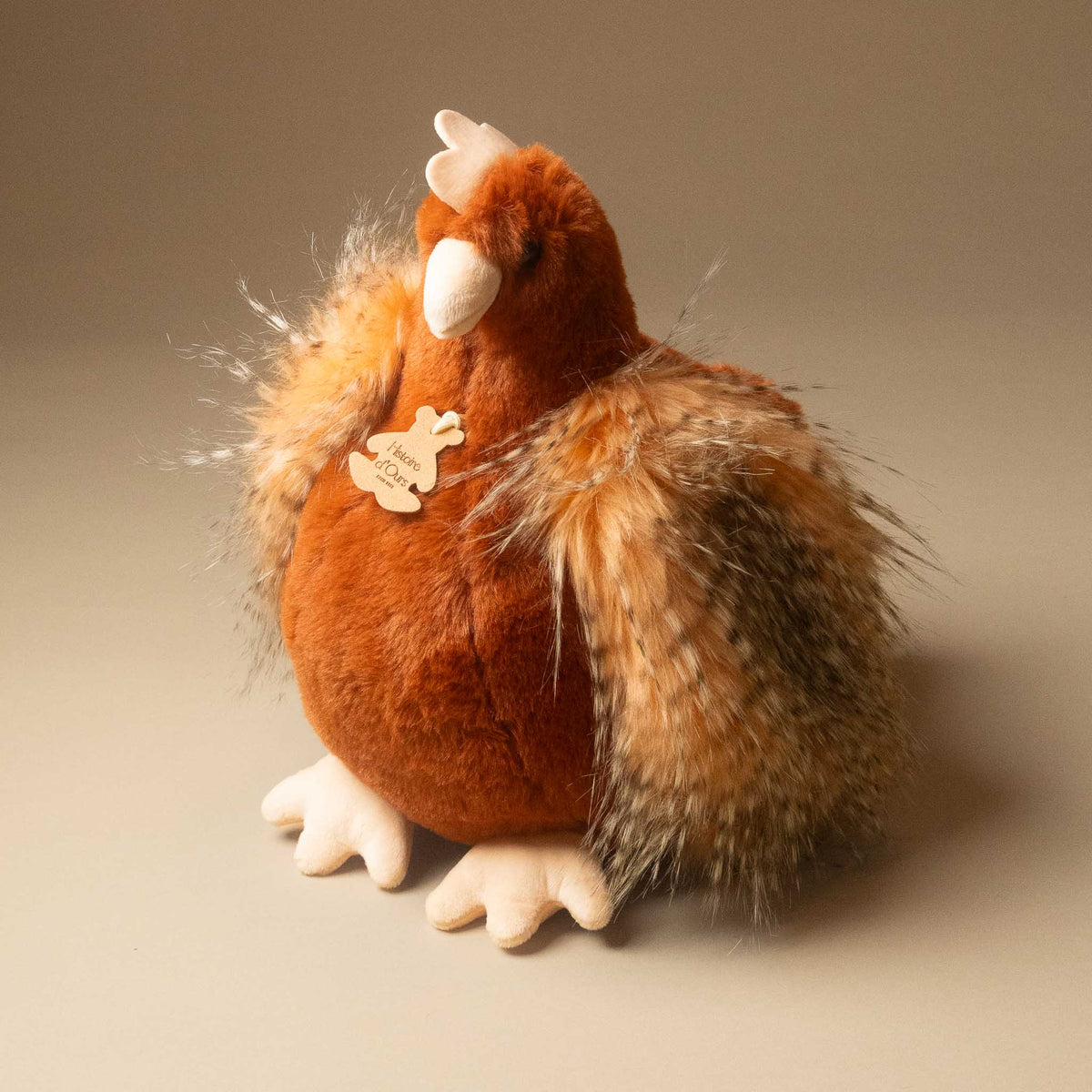 A plush floofy-chicken--her-golden-eggs--rouge with a round brown body, fluffy brown and black feathered wings, cream-colored beak and feet, and a small wooden tag on its side, photographed against a plain warm gray studio background.