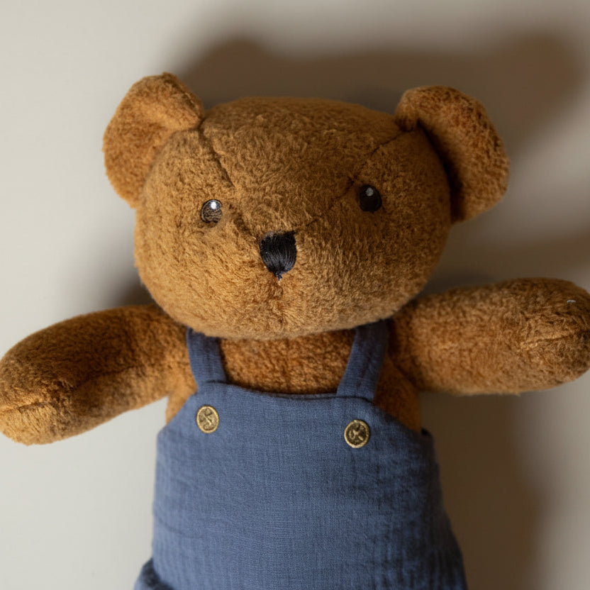gabin-the-Brown teddy bear wearing a blue overalls against a beige background