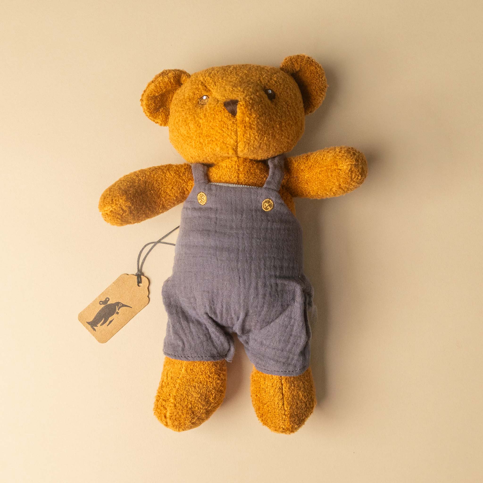gabin-the-bear wearing blue overalls with a tag on a beige background