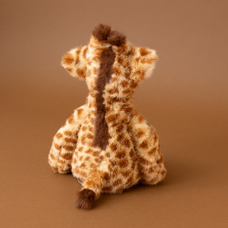 A plush giraffe toy shown from the back, featuring a textured brown and beige spotted pattern on a solid brown studio background.