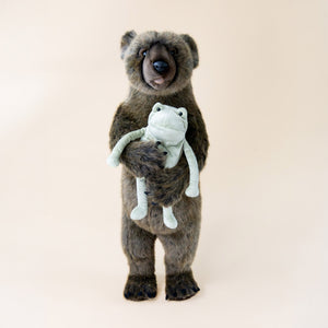 grizzly-bear-standing-small-with-wrapped-arms-and-bark-colored-fur-face-with-brown-snout-black-nose-bright-eyes-and-open-mouth-holding-frog-stuffed-animal