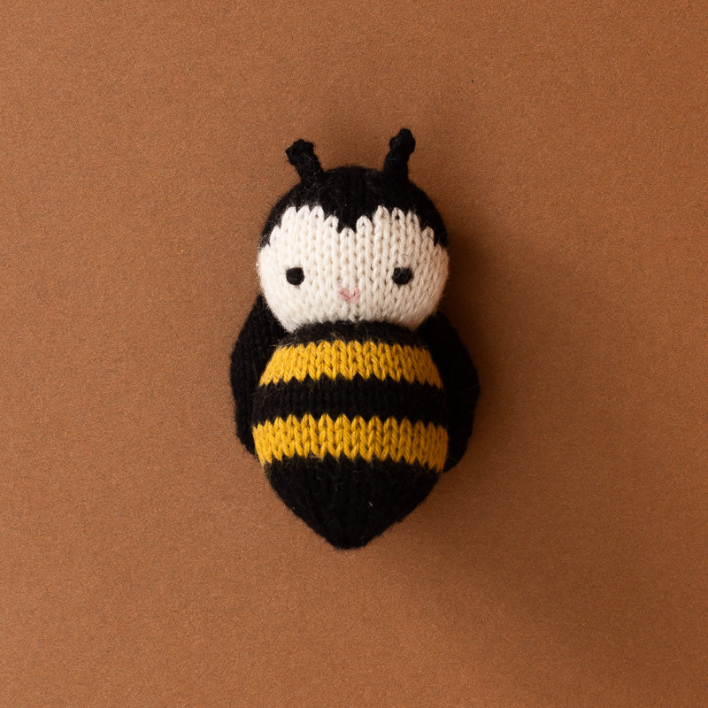 A hand-knit bumblebee plush toy or cushion, featuring a black and yellow striped body, a white face, and black antennae, set against a solid brown background.