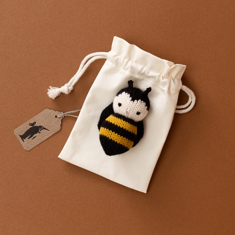 An off-white cotton drawstring pouch with a knitted bumblebee applique on the front, shown on a brown background with a small paper tag.