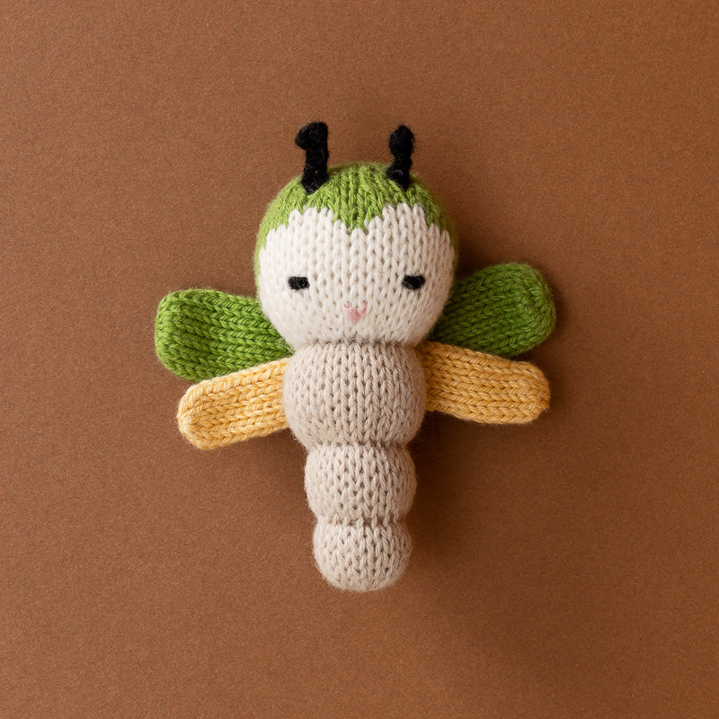 A hand-knitted toy dragonfly with a cream-colored body, green head and wings, and yellow wings, set against a solid brown background.