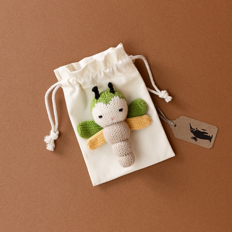 A small knitted dragonfly  plush toy with green wings and a beige body, placed on top of a white drawstring pouch. A small paper tag with a black fish logo is attached to the pouch.