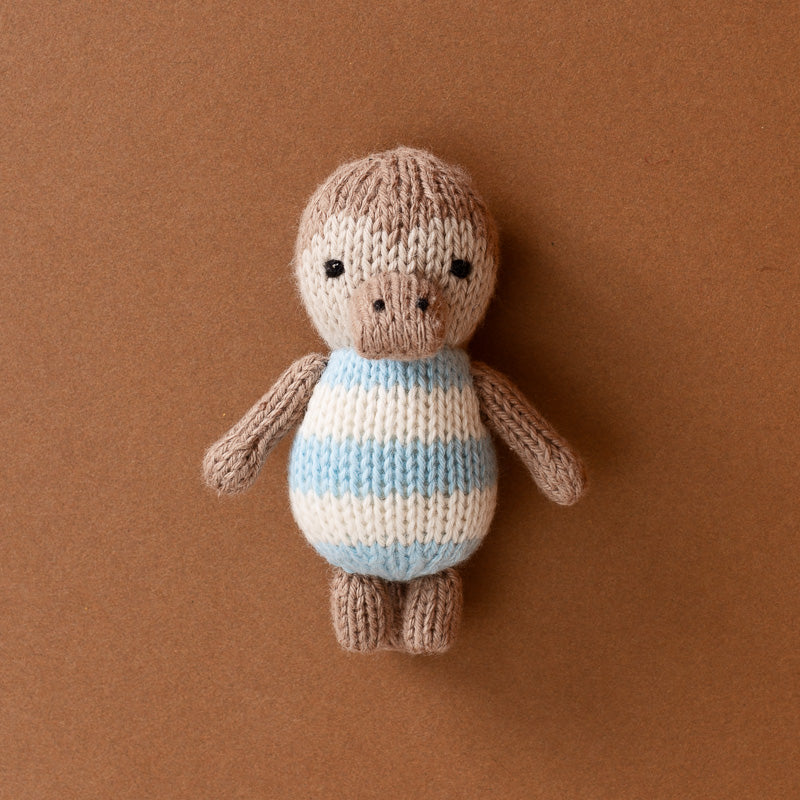 A small hand-knit-pocket-platypus   toy with a brown body, beige face and limbs, and a striped blue, white, and beige torso, centered on a solid brown background.