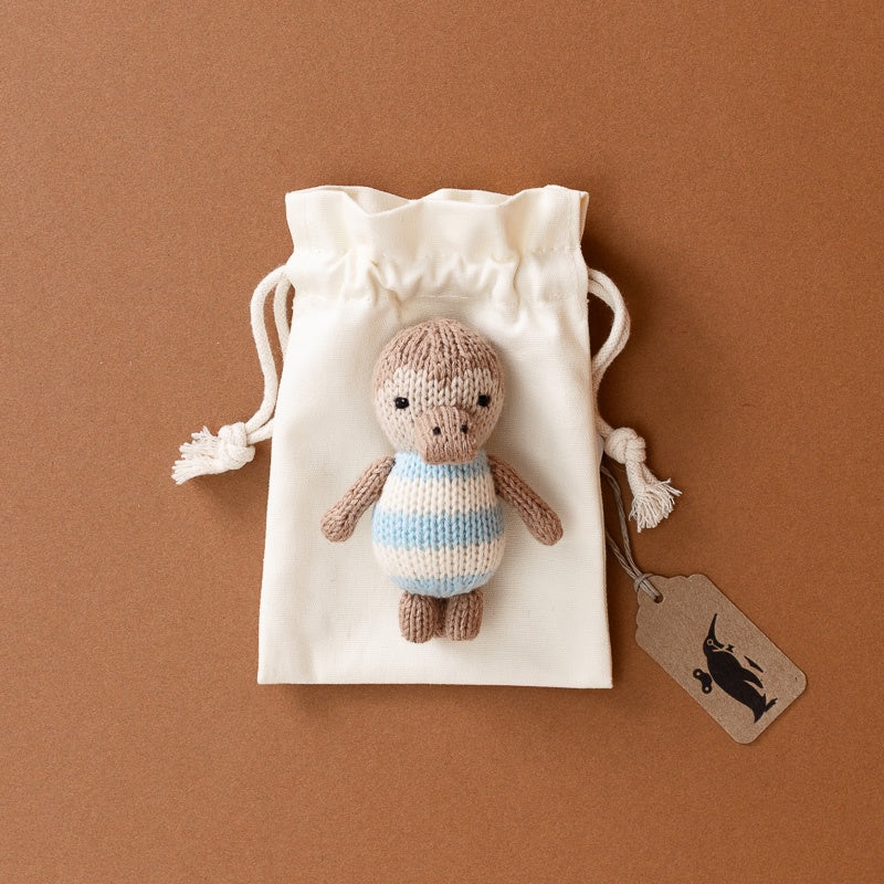 A small hand-knit-pocket-platypus toy with a brown knit body and a light blue and white striped shirt, displayed on top of a cream drawstring pouch. A brown paper hangtag with a black rabbit logo is attached to the side.