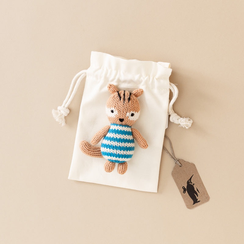 A small knitted squirrel plush toy wearing a blue and white striped shirt, placed on a white drawstring pouch with a small brown tag attached, photographed on a warm beige background.