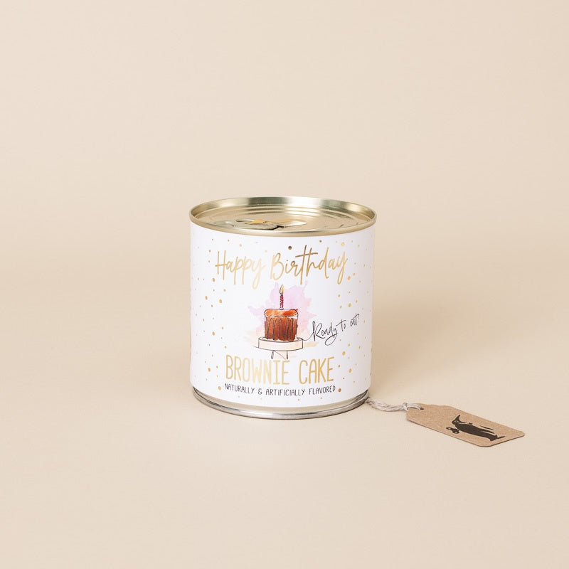 A white candle tin with a gold lid, featuring a 'Happy Birthday' design with a brownie cake illustration and the text 'BROWNIE CAKE'. A small kraft paper hangtag is attached to the side, set against a solid beige background.