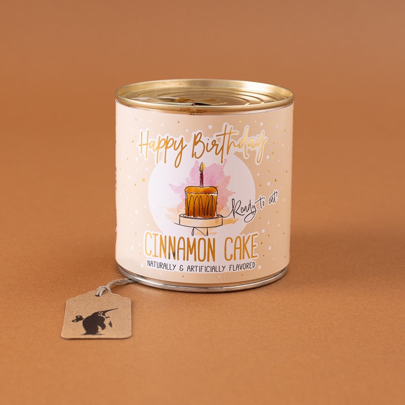 A novelty candle in a tin canister with a gold lid, featuring a 'Happy Birthday' and 'Cinnamon Cake' birthday candle design on a warm beige label. A small paper tag with a black rat illustration hangs from the side.