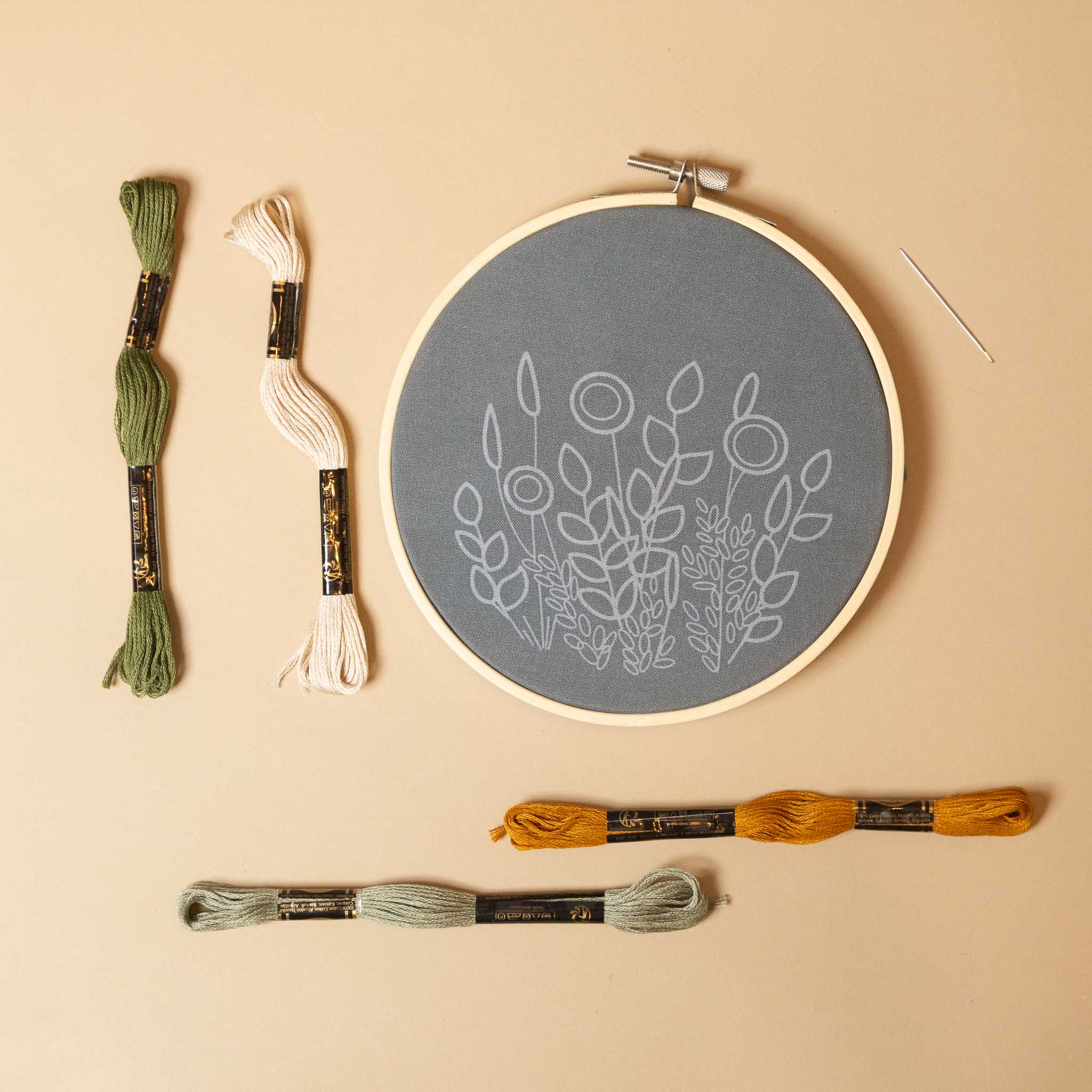 hawthorne-embroidery-kit-green-and-mustard Embroidery hoop with floral pattern and embroidery threads on a beige background