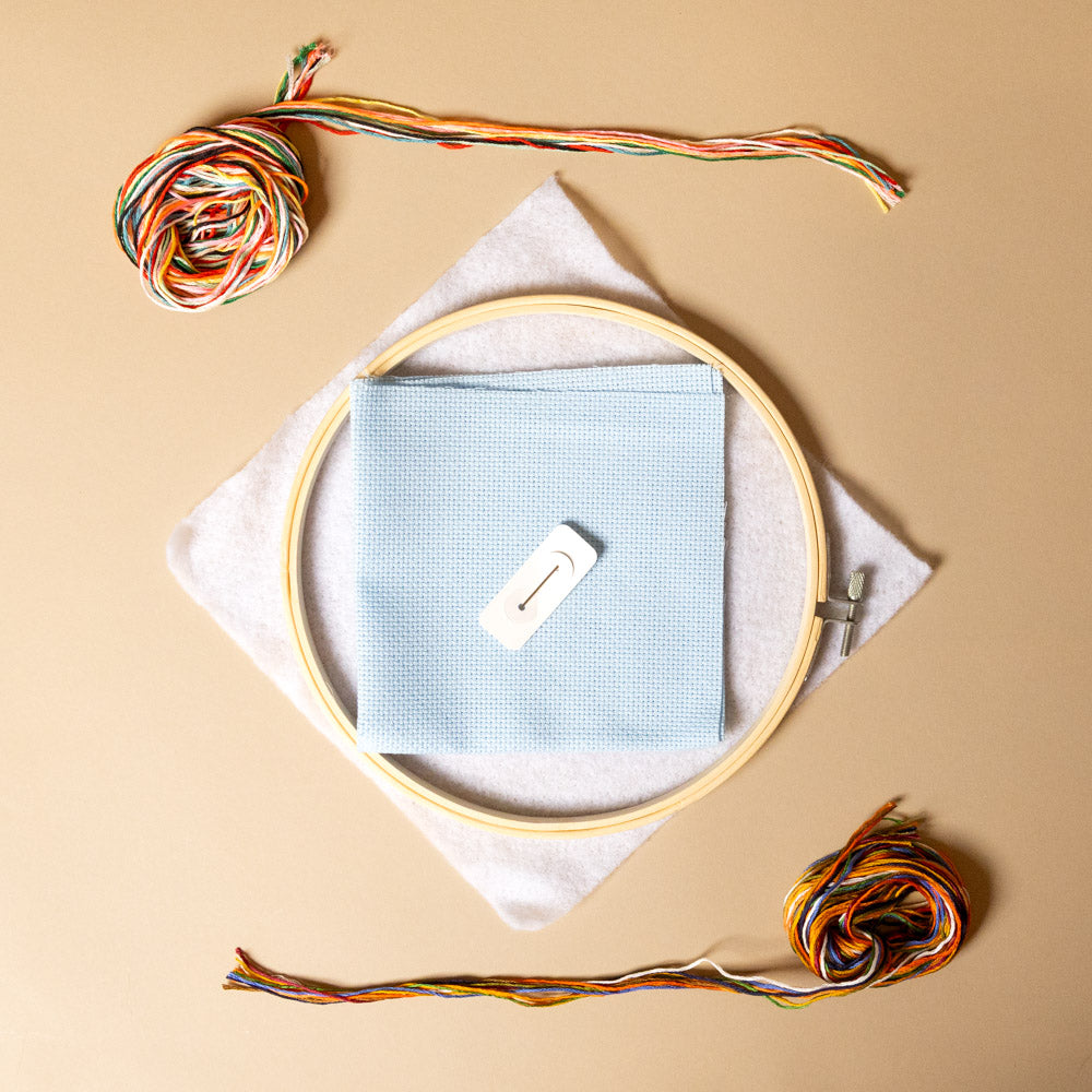  holiday-cross-stitch-kit--beginner Embroidery hoop with light blue fabric, wooden clip, and colorful thread on a beige background