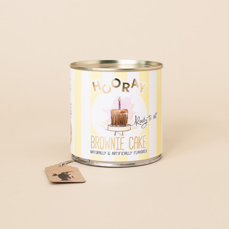 A tin of Hooray Brownie Cake on a warm beige background, with a small brownie tag attached to the side. The label features a yellow and white striped design with an illustration of a brownie cake with a single candle.