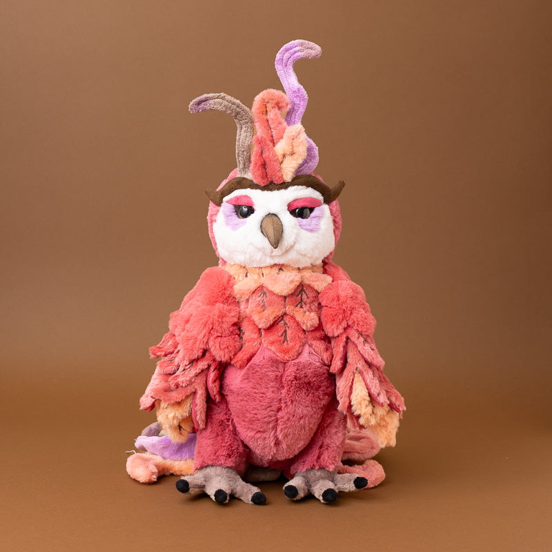 A plush jinzi-the-rising-phoenix toy with a pink and coral feathered body, a white face, and a tall multicolored crest, posed against a solid brown studio background.