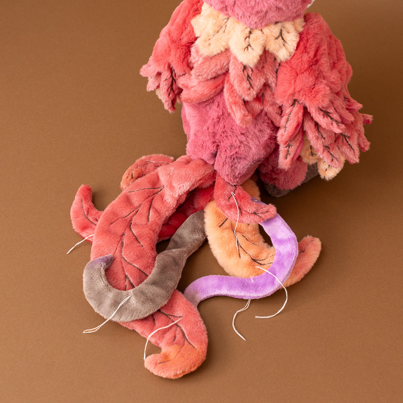 A close-up of a handmade plush jinzi-the-rising-phoenix toy or stuffed animal body made from fuzzy fabric strips in pink, coral, peach, purple, and gray, arranged on a solid brown background.