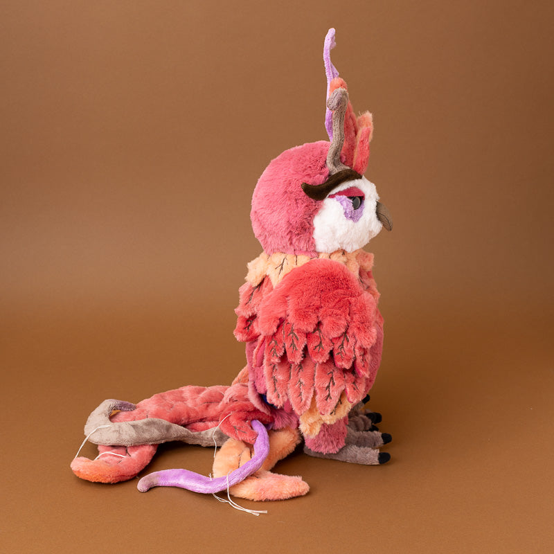 A plush toy jinzi-the-rising-phoenix bird with layered pink, coral, and peach feathers, a white face patch, and purple and gray accents, posed against a solid brown studio background.
