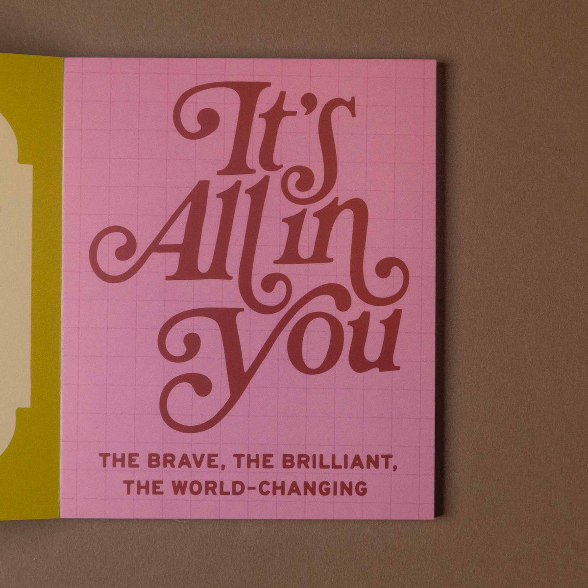 A square pink card with a subtle grid pattern and large cursive text reading 'It's All in You,' with smaller text below, photographed against a brown background.