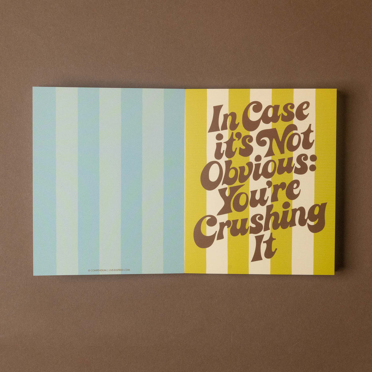 A square art greeting card with a split design, featuring light blue and white vertical stripes on the left and yellow and white vertical stripes on the right with the text 'In Case it's Not Obvious: You're Crushing It' in brown retro lettering, displayed on a solid brown background.