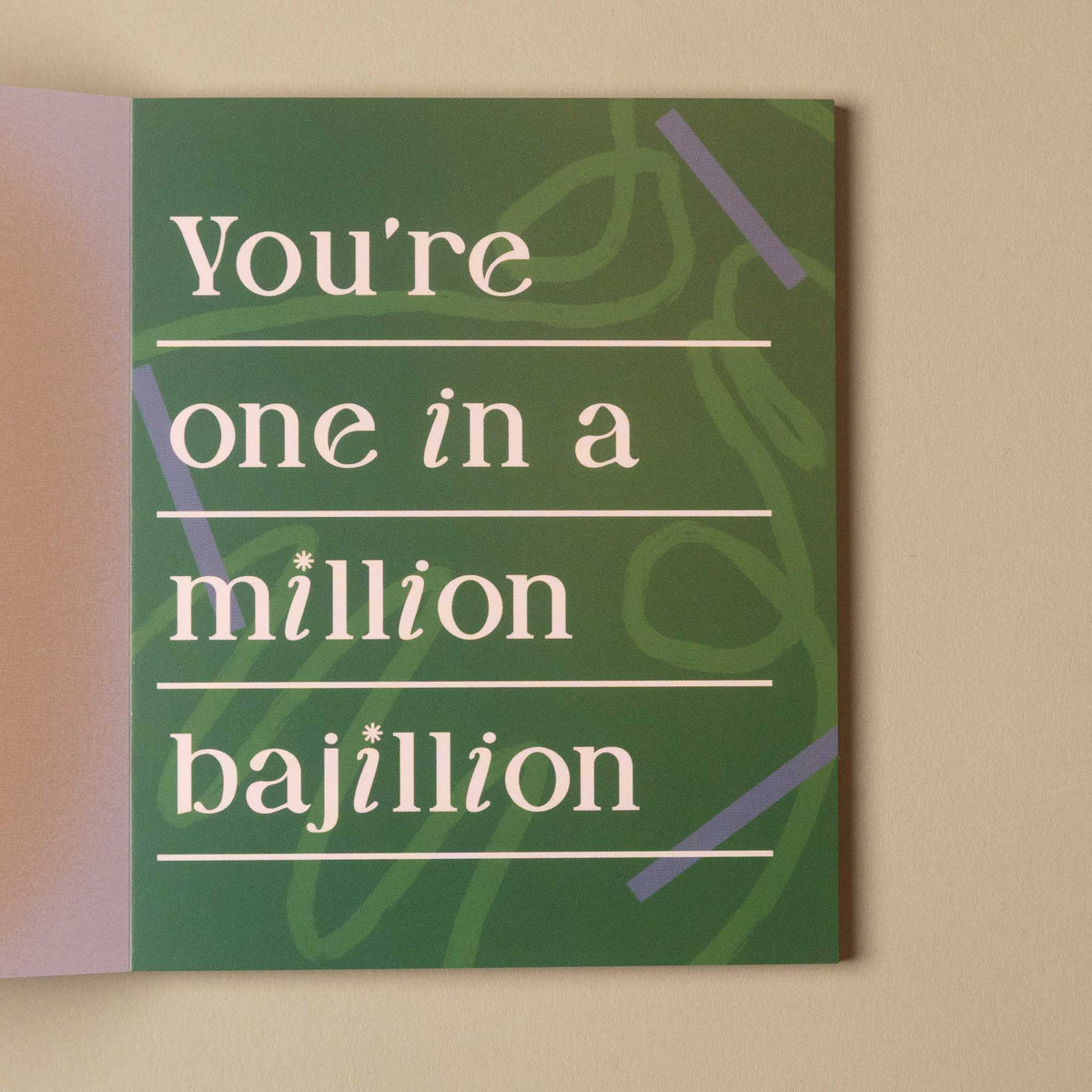 A square greeting card with a green background and white text that reads 'You're one in a million bajillion' against a beige wall.