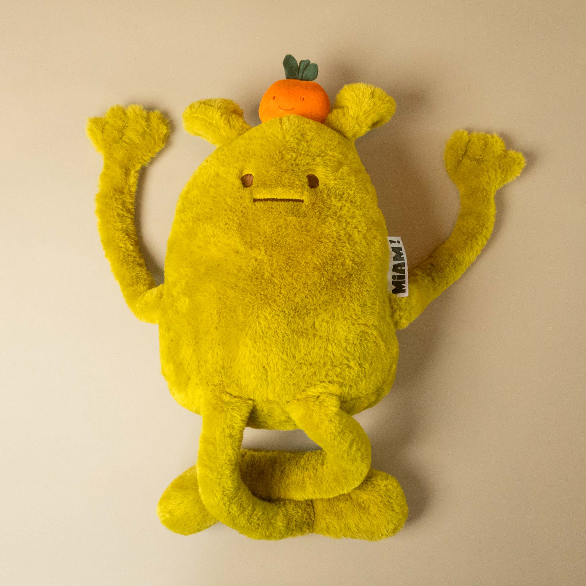 A yellow laurent-jade-grande plush toy with long arms and legs, a small orange fruit on its head on a plain beige studio background.
