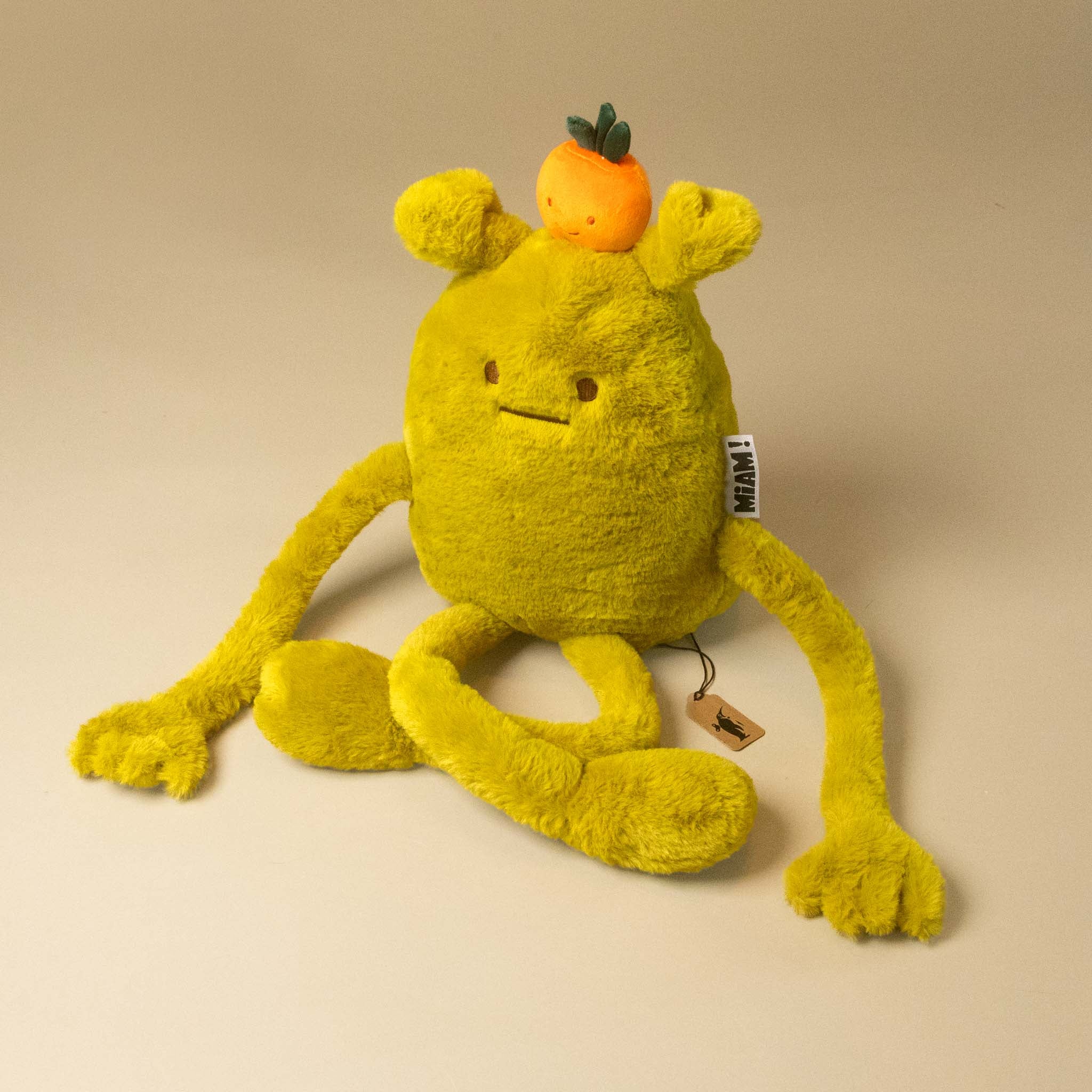 A yellow laurent-jade-grande plush toy with long arms and legs, a small orange fruit on its head on a plain beige studio background.