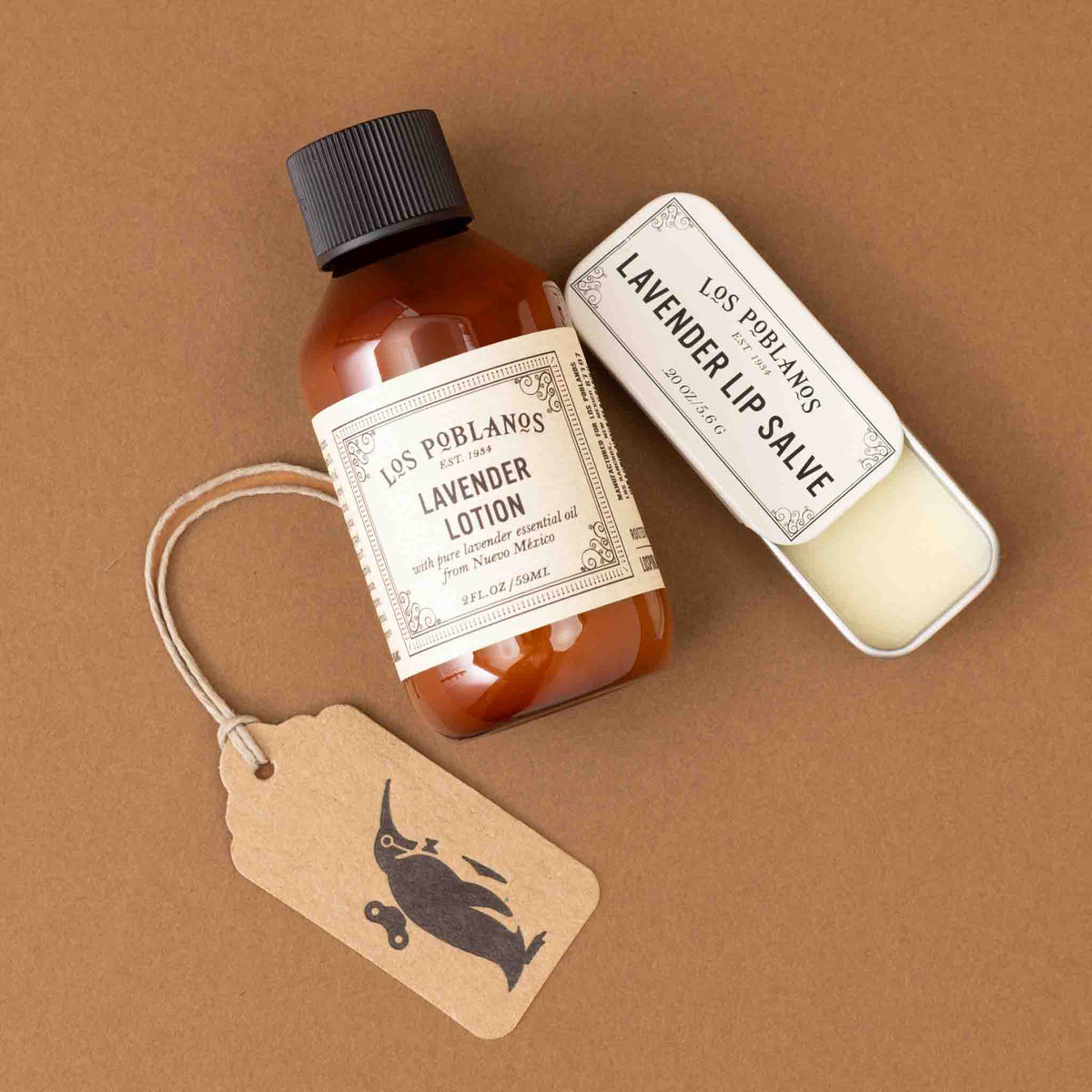 A bottle of Los Portales Lavender Lotion and a tin of Lavender Lip Salve arranged on a brown background with a kraft paper hangtag featuring a black bird logo.