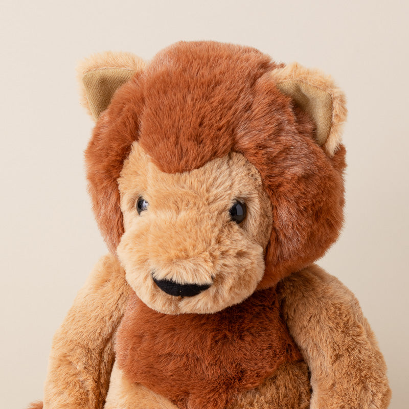 A plush lion toy with a warm brown mane, tan face and paws, and small black eyes and nose, photographed against a plain light beige background.