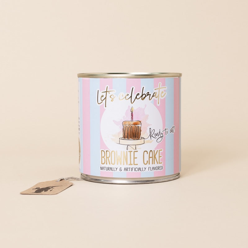 A cylindrical lets-celebrate-cancake-brownie-cake with a pastel pink and blue striped label featuring a brownie cake illustration and the text 'let's celebrate Ready to art BROWNIE CAKE'. A small kraft paper tag hangs from the side.
