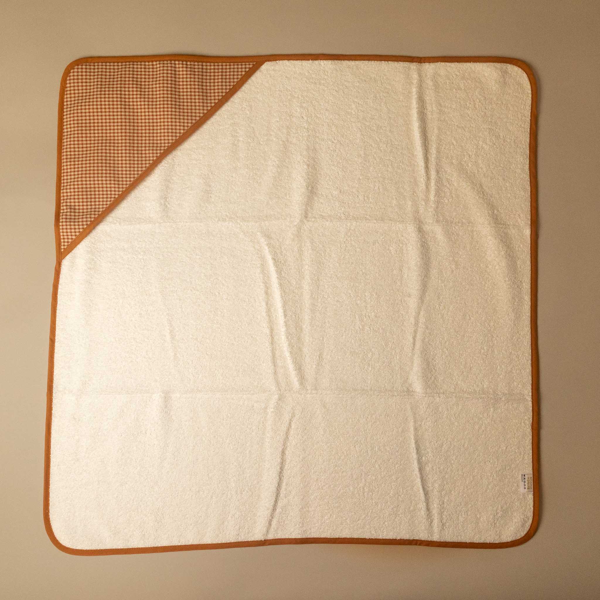 A square baby towel with a cream-colored terry cloth body, a brown gingham checkered hood corner, and a brown border, laid flat on a neutral beige background.