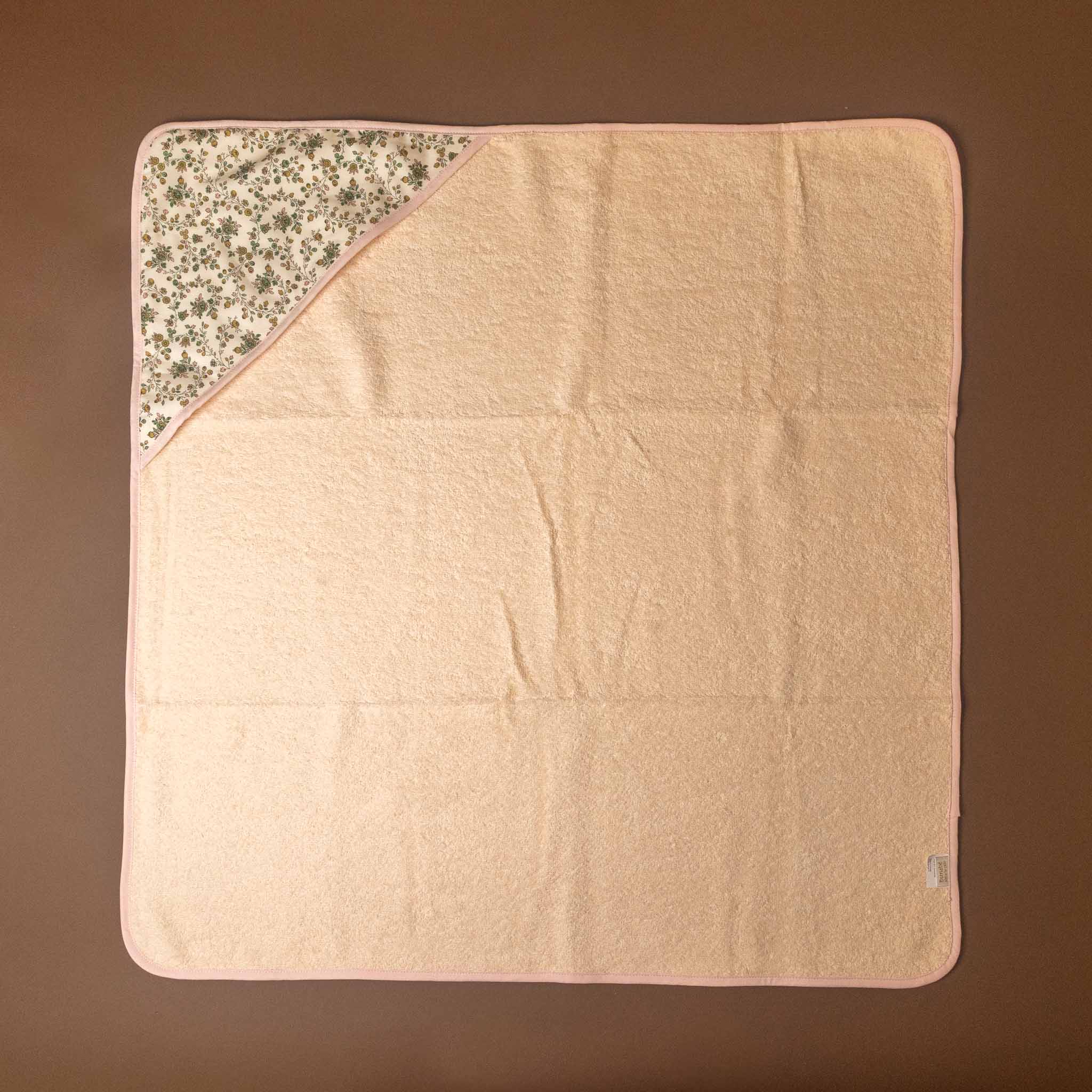 A square baby blanket or towel in pale peach-pink, laid flat on a brown background. One corner is folded over to reveal a floral patterned hood in beige, white, and muted green.