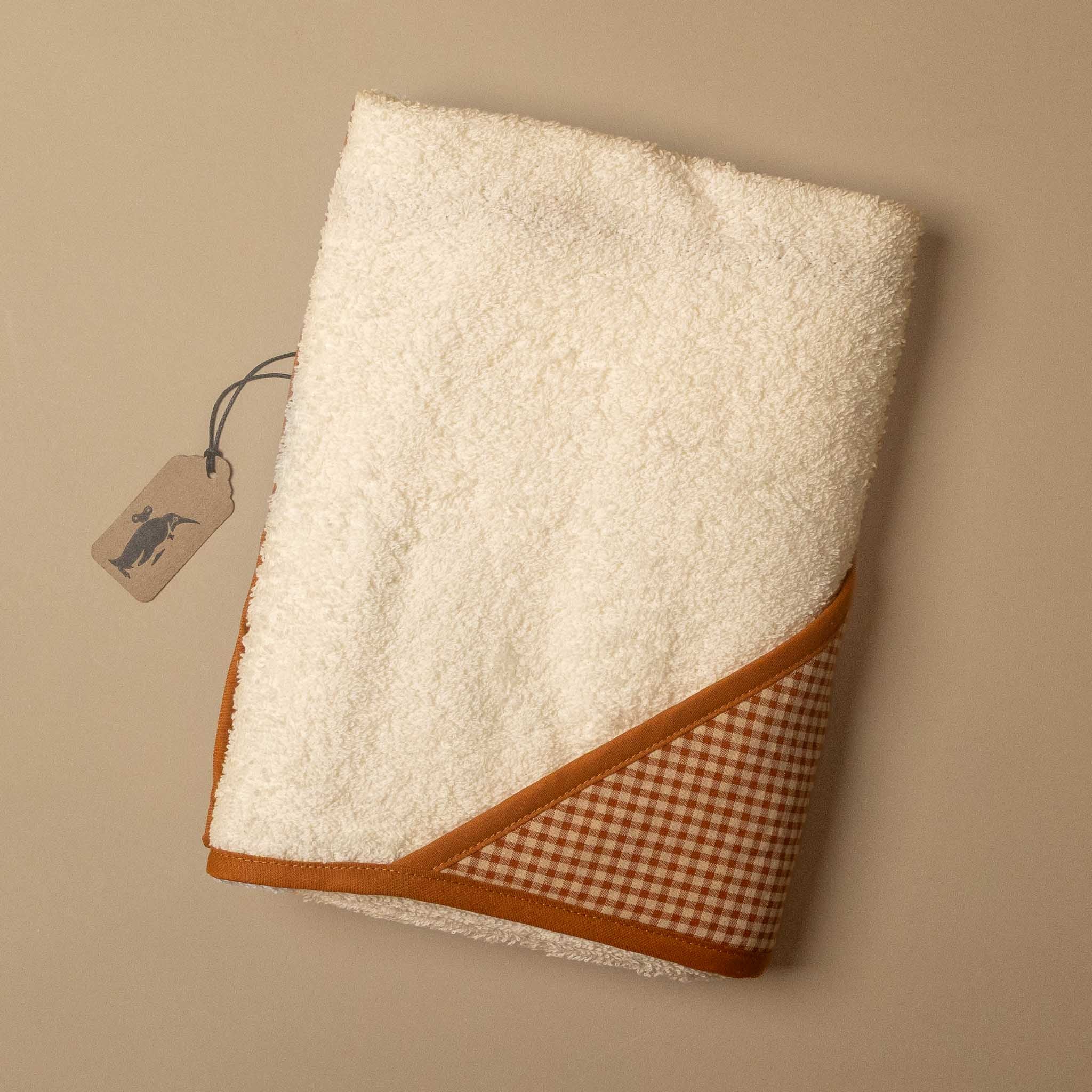 A folded cream-colored hooded towel with a brown gingham check pattern and a brown trim, shown on a neutral beige background with a small paper tag attached.