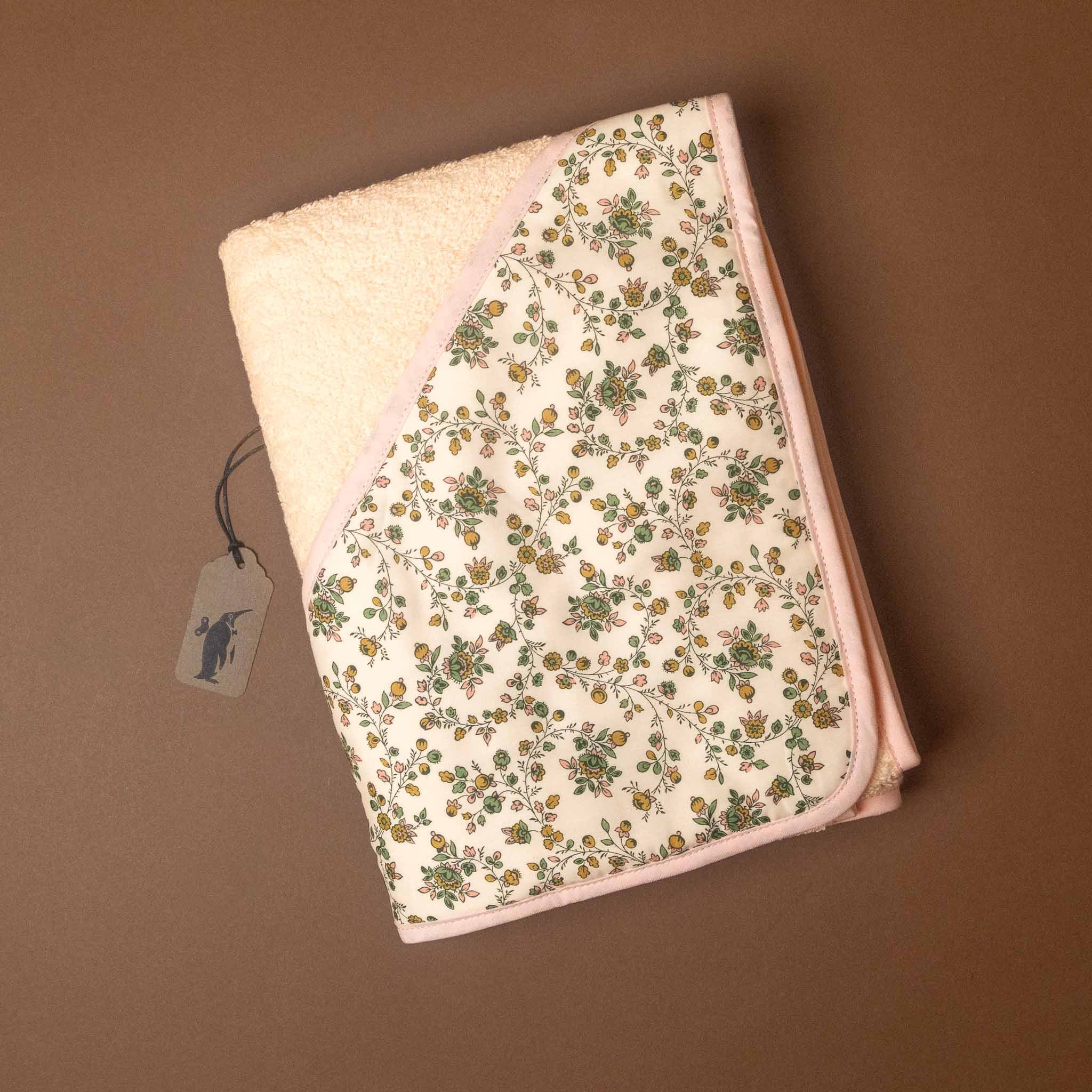 A folded baby towel with a cream-colored plush hood and a front panel featuring a delicate green and yellow floral pattern on a cream background, finished with light pink trim. A small brand tag is attached on the left, set against a solid brown background.