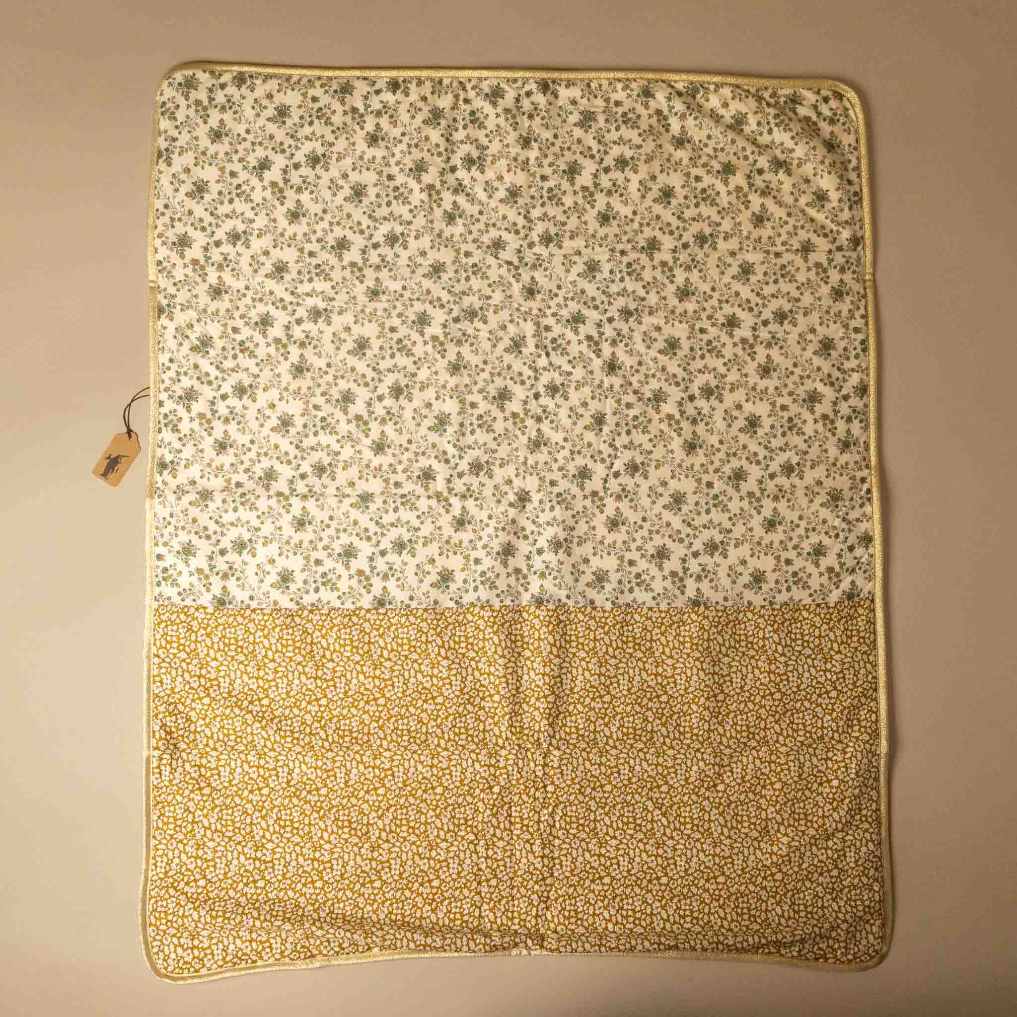 A square fabric blanket or quilt laid flat on a neutral beige background, featuring a two-tone floral pattern. The top half has a small green floral print on an off-white background, and the bottom half has a dense small yellow floral print on a warm beige background.