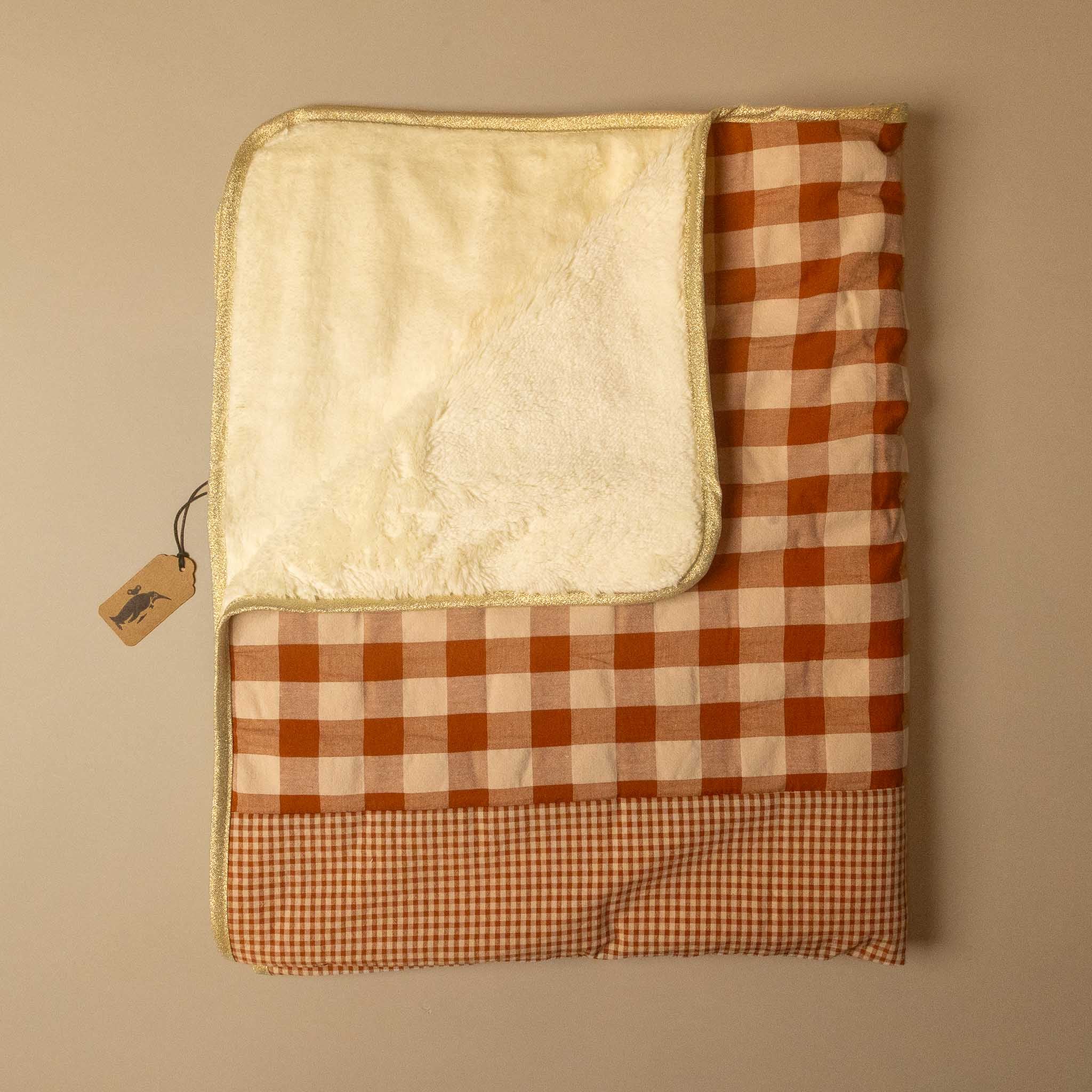 A folded quilted blanket with a warm cream sherpa lining and a rust-orange and cream gingham pattern, displayed on a neutral beige background with a small paper tag attached.