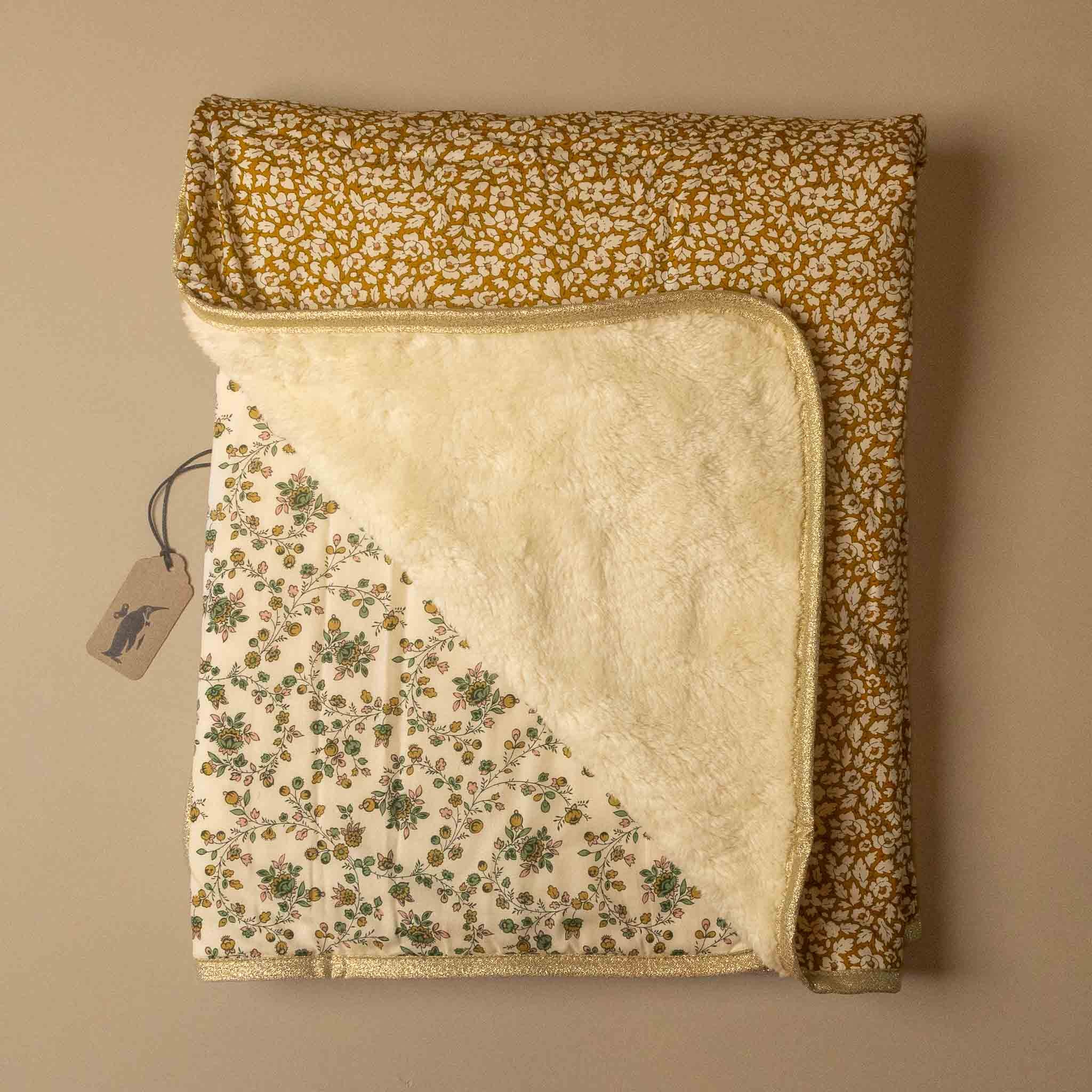 A folded baby blanket with a mustard yellow and white speckled pattern on one side, a cream sherpa fleece lining on the reverse, and a green and cream floral print on the corner. A small paper tag is attached.