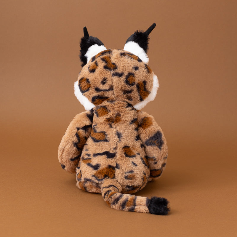 A plush toy lynx shown from the back, featuring a brown and black spotted pattern, white accents around the face, and black-tipped ears and tail, set against a solid brown background.