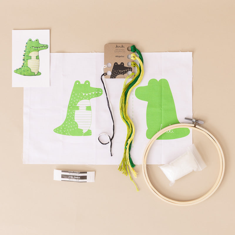 Embroidery kit components laid out on a beige background, including a white fabric panel with green alligator embroidery designs, a wooden hoop, thread spools, a small card, and a tag.