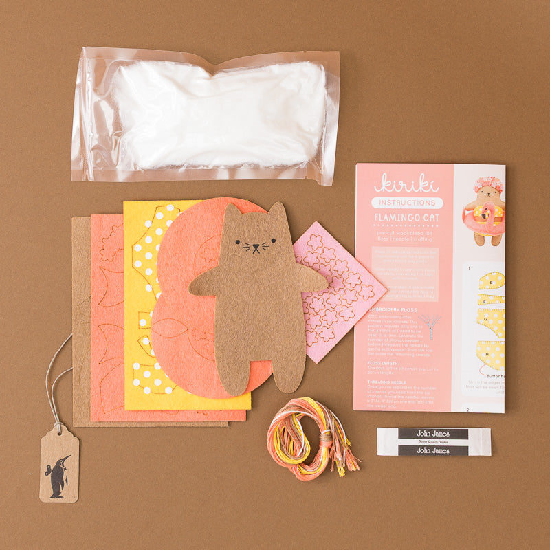 A flat-lay of a craft kit on a brown background, including a clear pouch with white fabric, patterned paper sheets, a brown felt cat cutout, pink patterned paper, a printed instruction booklet titled 'Flamingo Cat', thread, and two small labels.