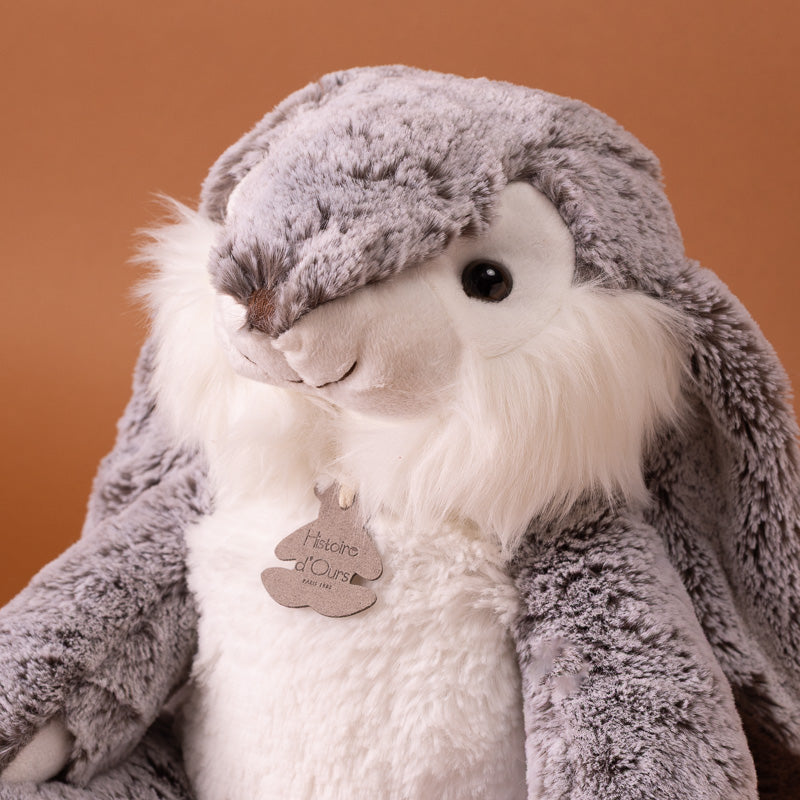 A plush stuffed bunny toy with long floppy ears, sitting upright against a solid warm brown background. The bunny has gray and white fur