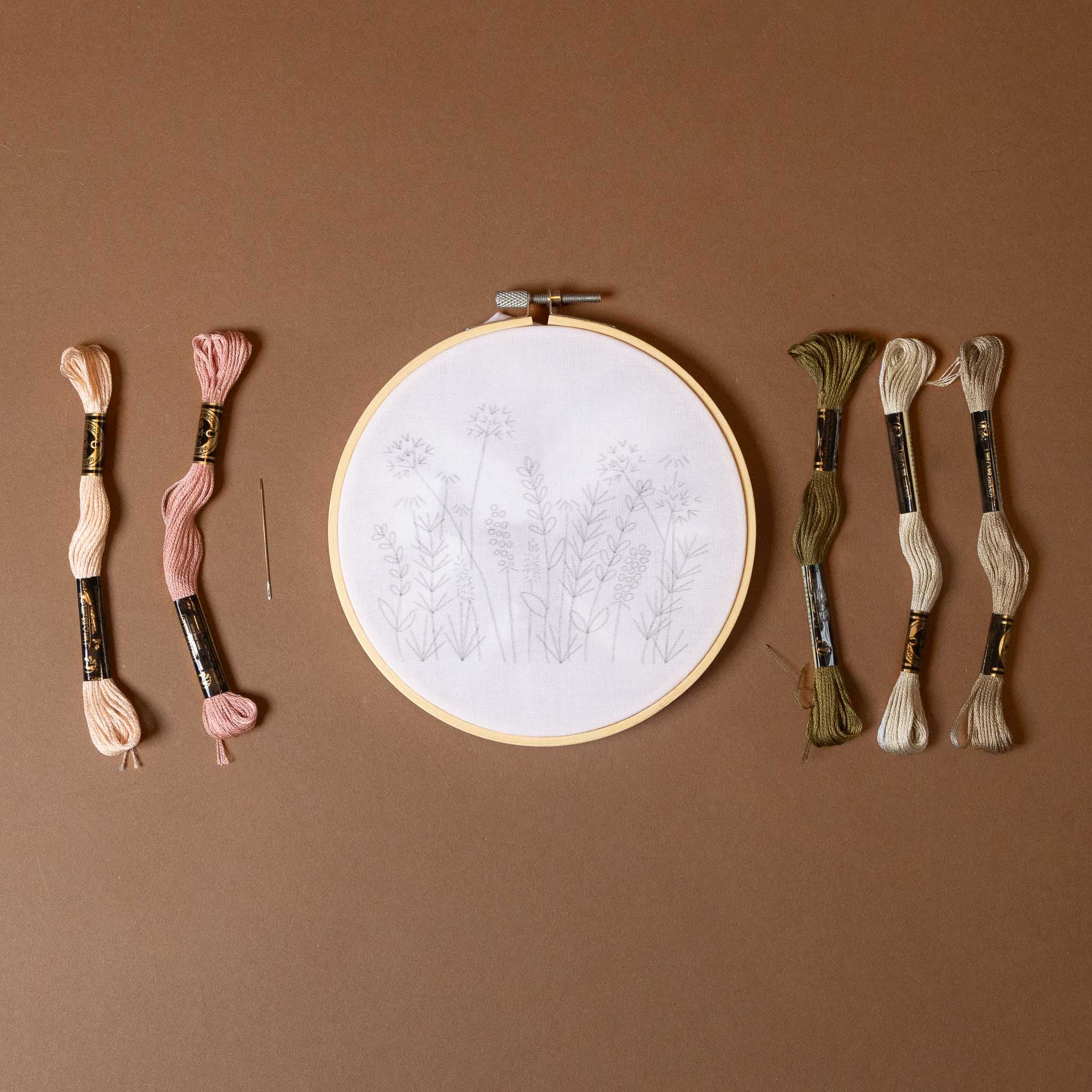 meadow-embroidery-kit-blush-and-oliv Embroidery hoop with white fabric and assorted embroidery threads on a brown background