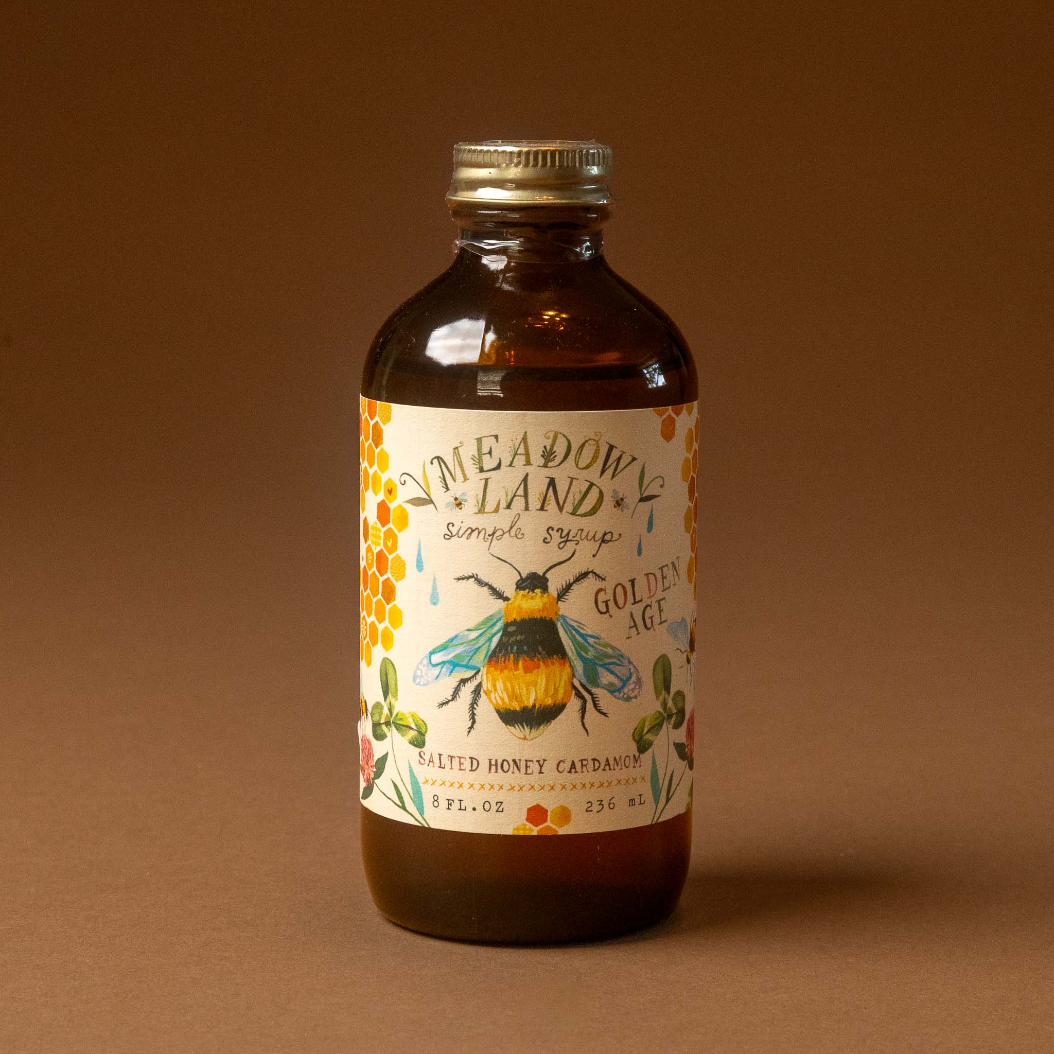 An amber glass bottle of meadowland-simple-syrup-golden-age with a gold cap and a detailed bee label, set against a solid brown background.