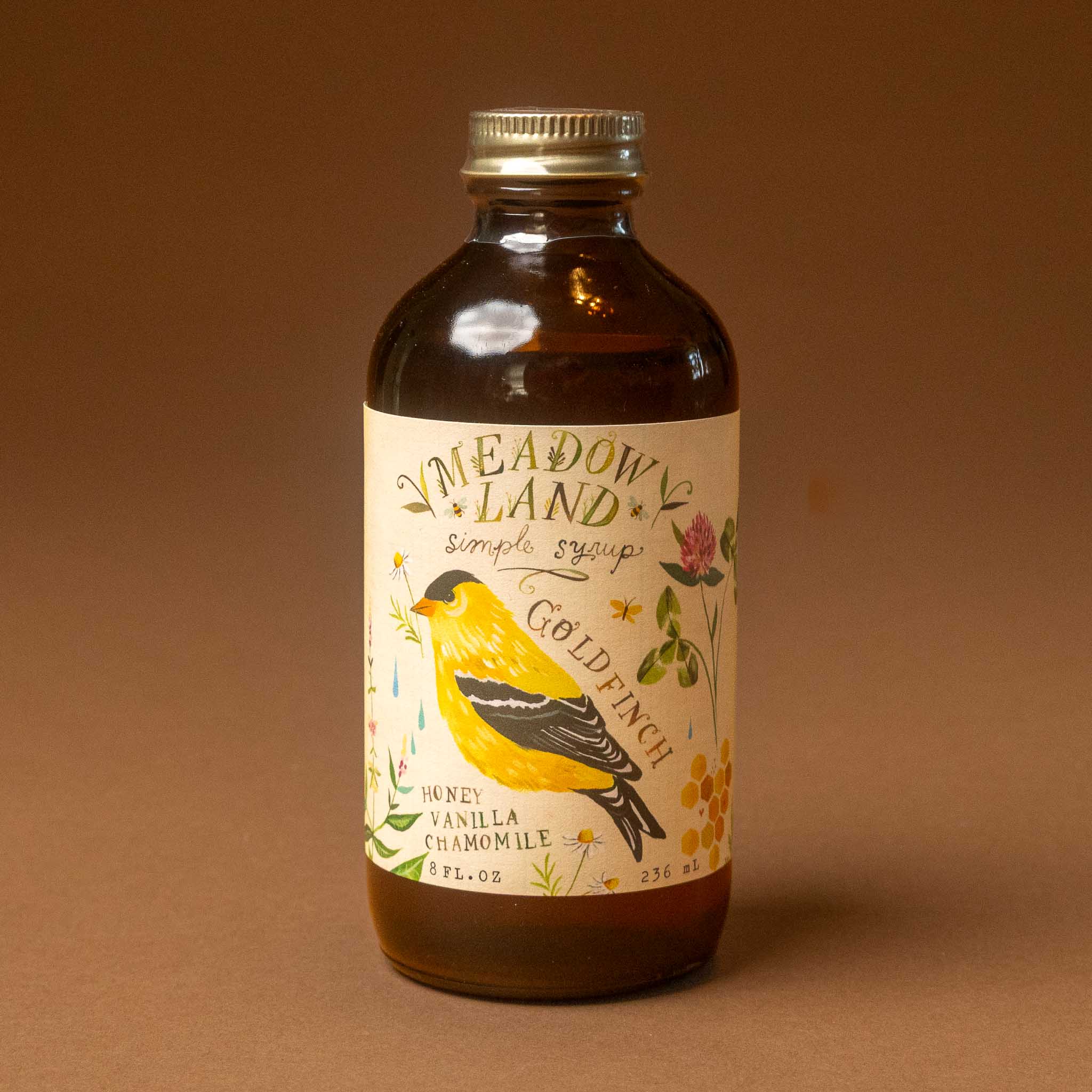 A bottle of Meadow's Land Gold Finch Simple Syrup against a solid brown background. The product is in an amber glass bottle with a gold cap and a cream-colored label featuring a yellow and black gold finch illustration.