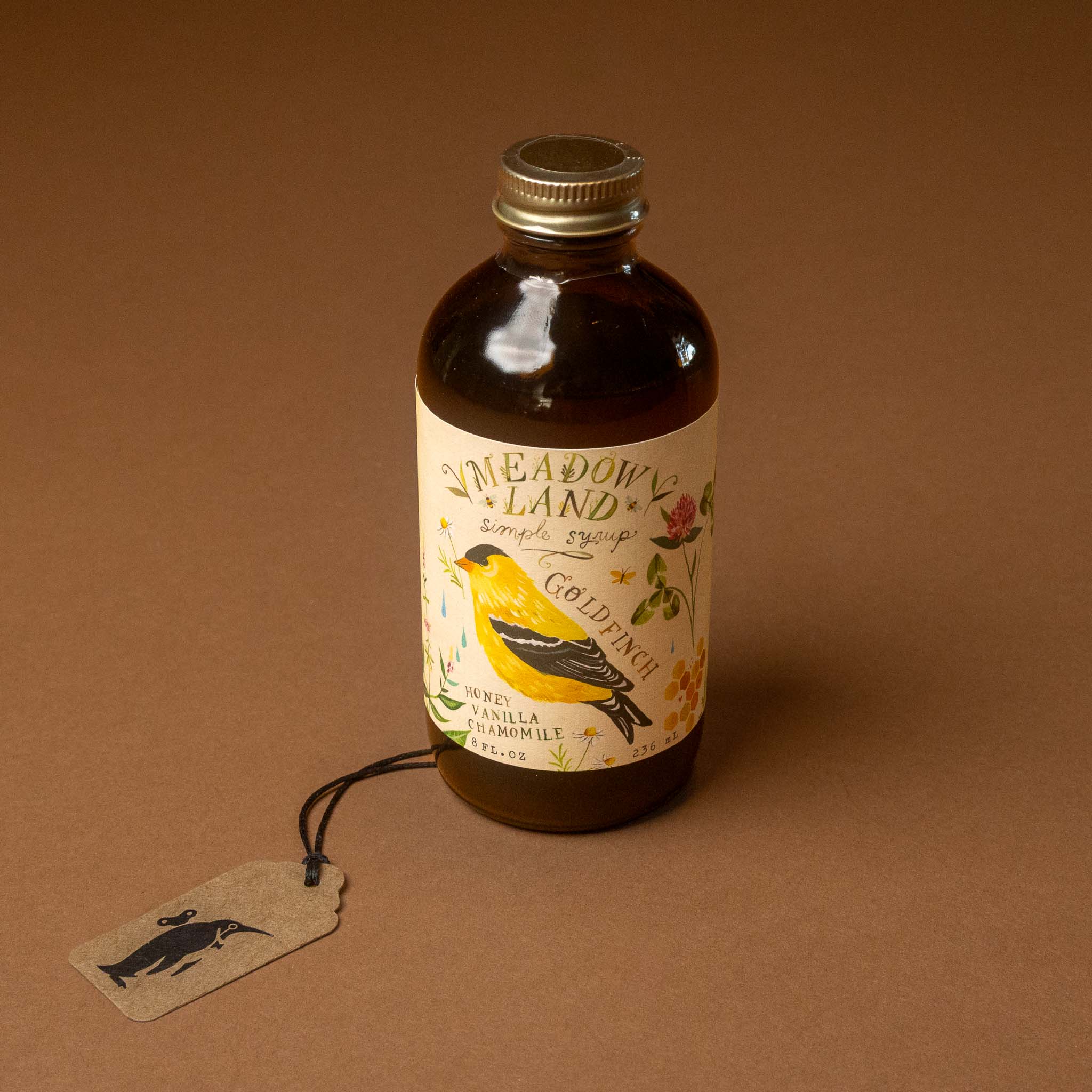 A bottle of Meadow Land Goldranch vanilla vanilla-flavored syrup with a yellow bird illustration on the label, set against a solid brown background with a hanging paper tag.