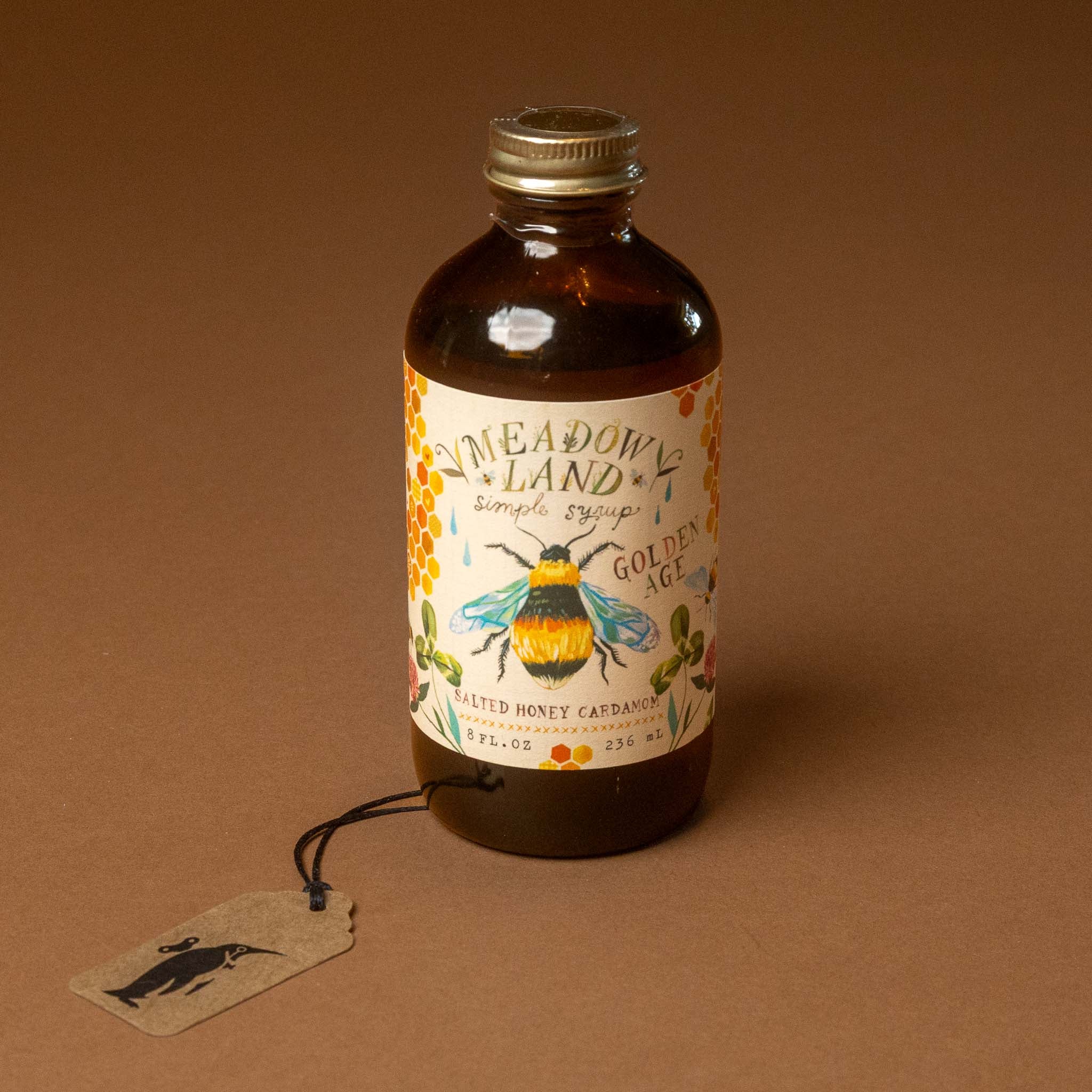 An amber glass bottle of meadowland-simple-syrup-golden-age with a cream label featuring a bumblebee illustration, set against a solid brown background with a hanging paper tag.
