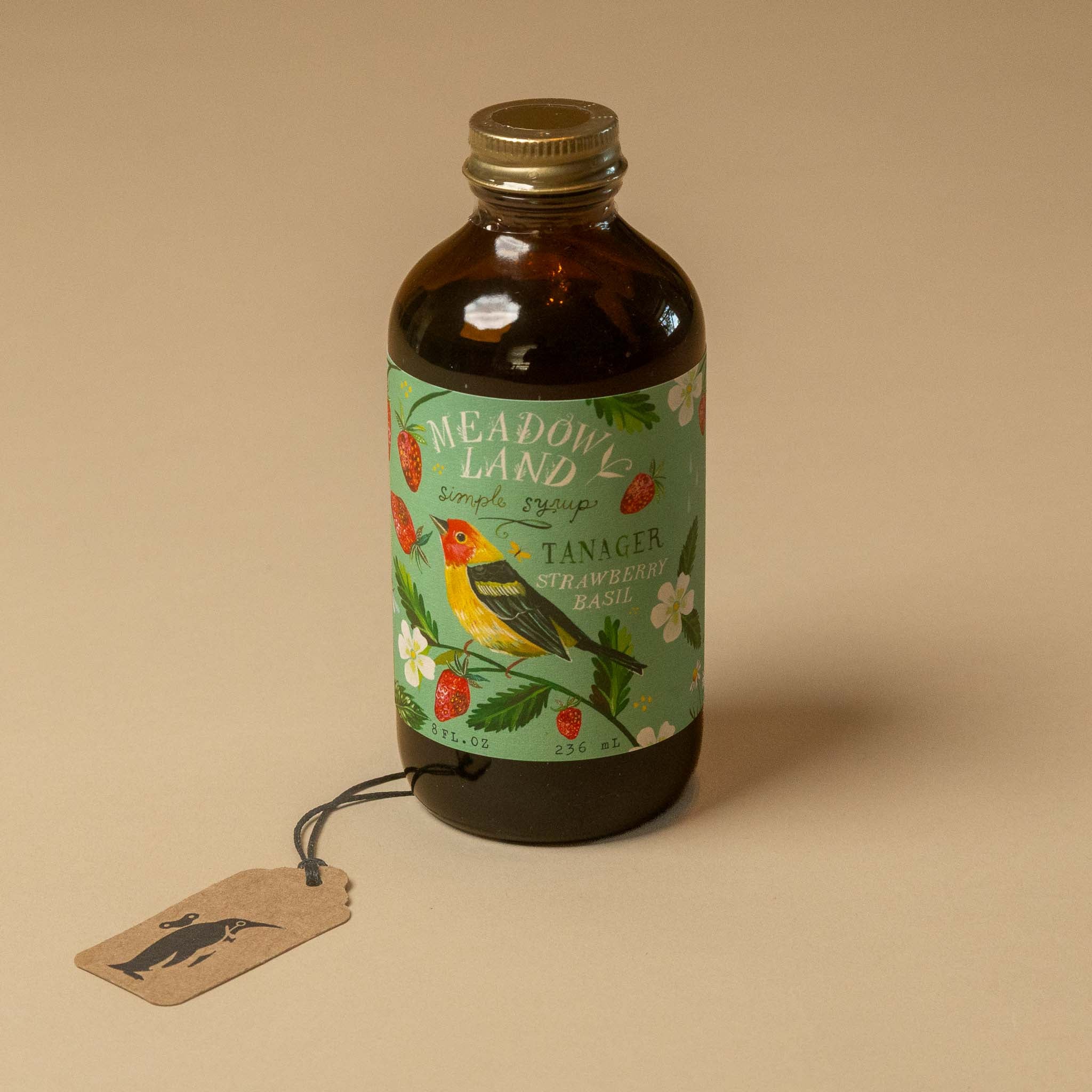 An amber glass bottle of meadowland-simple-syrup-tanager with a detailed green label featuring a yellow-breasted bird and strawberries, shown on a beige background with a hanging kraft paper tag.