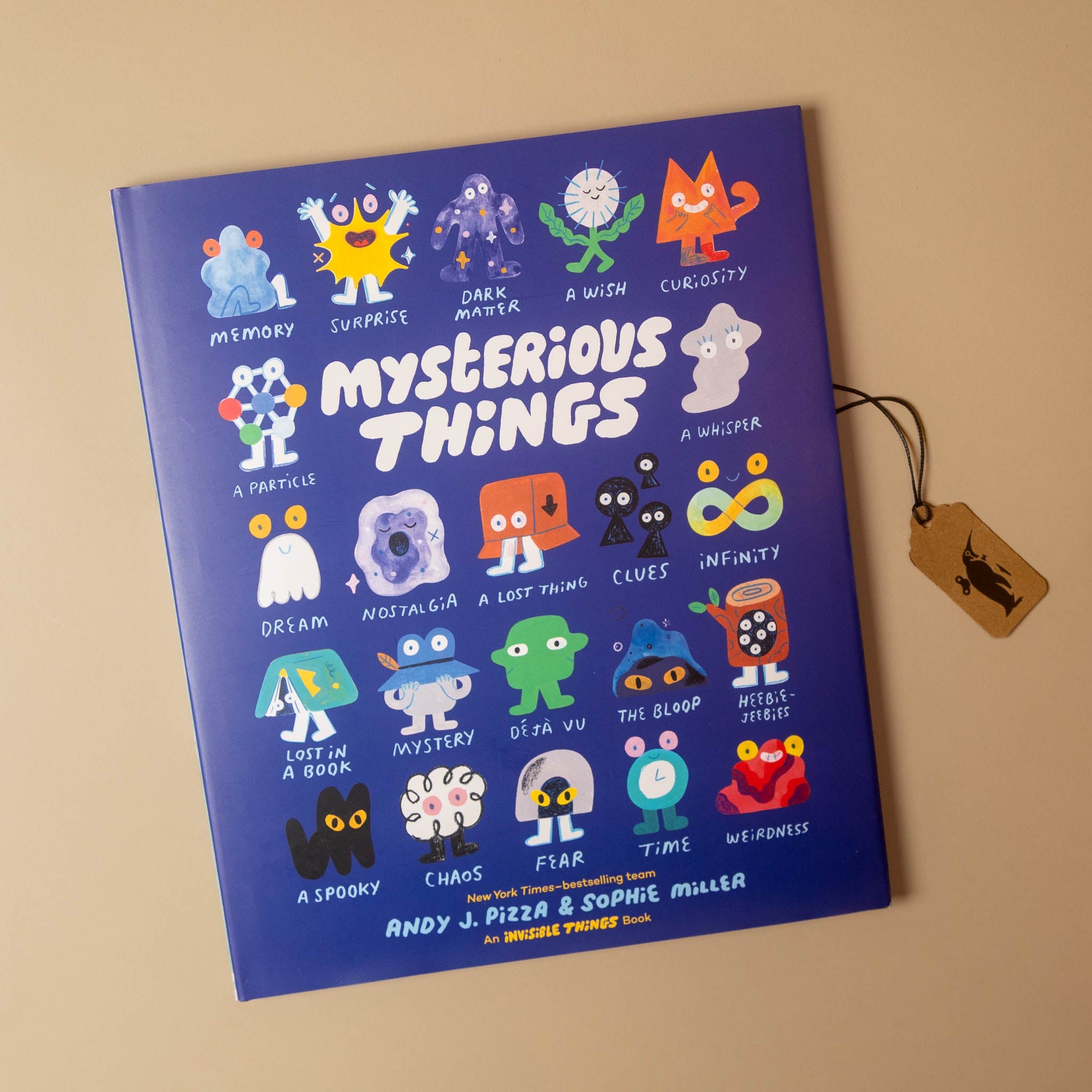 The book 'Mysterious Things' by Andy J. Pizza and Sophie Muller, featuring a vibrant blue cover illustrated with various colorful cartoon characters and titles, displayed on a warm beige background next to a wooden tag.