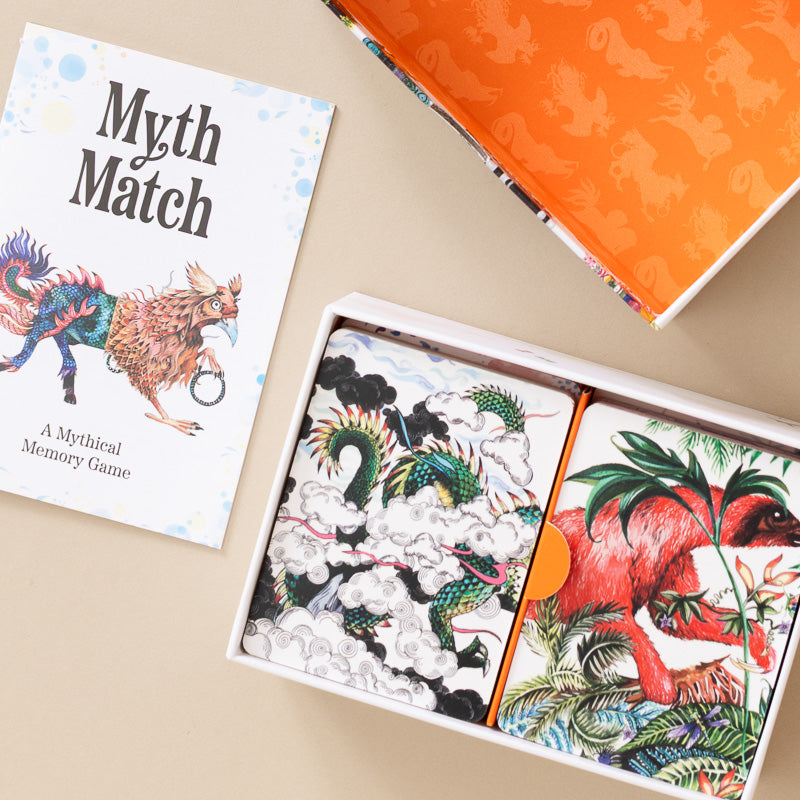 Myth Match: A Mythical Memory Game shown in its packaging, featuring a white card with a colorful creature illustration, an open box with illustrated card backs depicting a green dragon and a red dinosaur, and an orange patterned inner lid on a beige background.