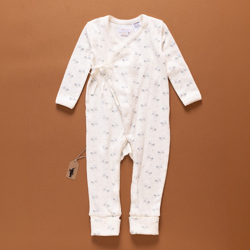 A baby's long-sleeve footless one-piece romper in cream fleece with a small blue floral print, laid flat on a solid brown background. The romper features a wrap-style front with a tie closure and a hangtag.