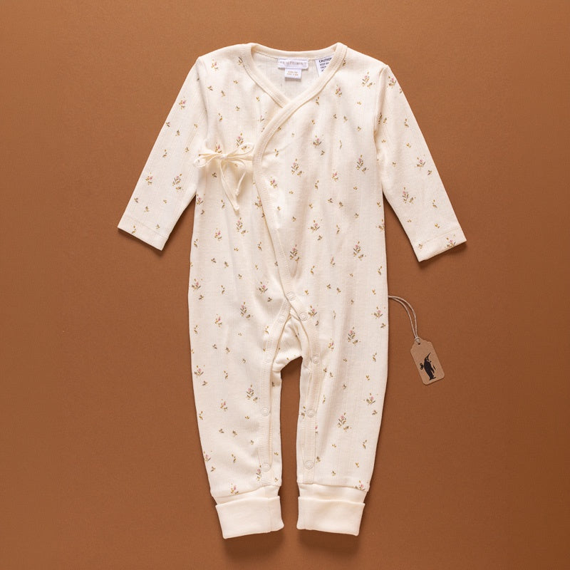 A cream-colored long-sleeve baby romper with a tiny floral print, laid flat on a warm brown background. The romper features a wrap-style front with a tie closure and a small bow, along with ribbed cuffs and hem.